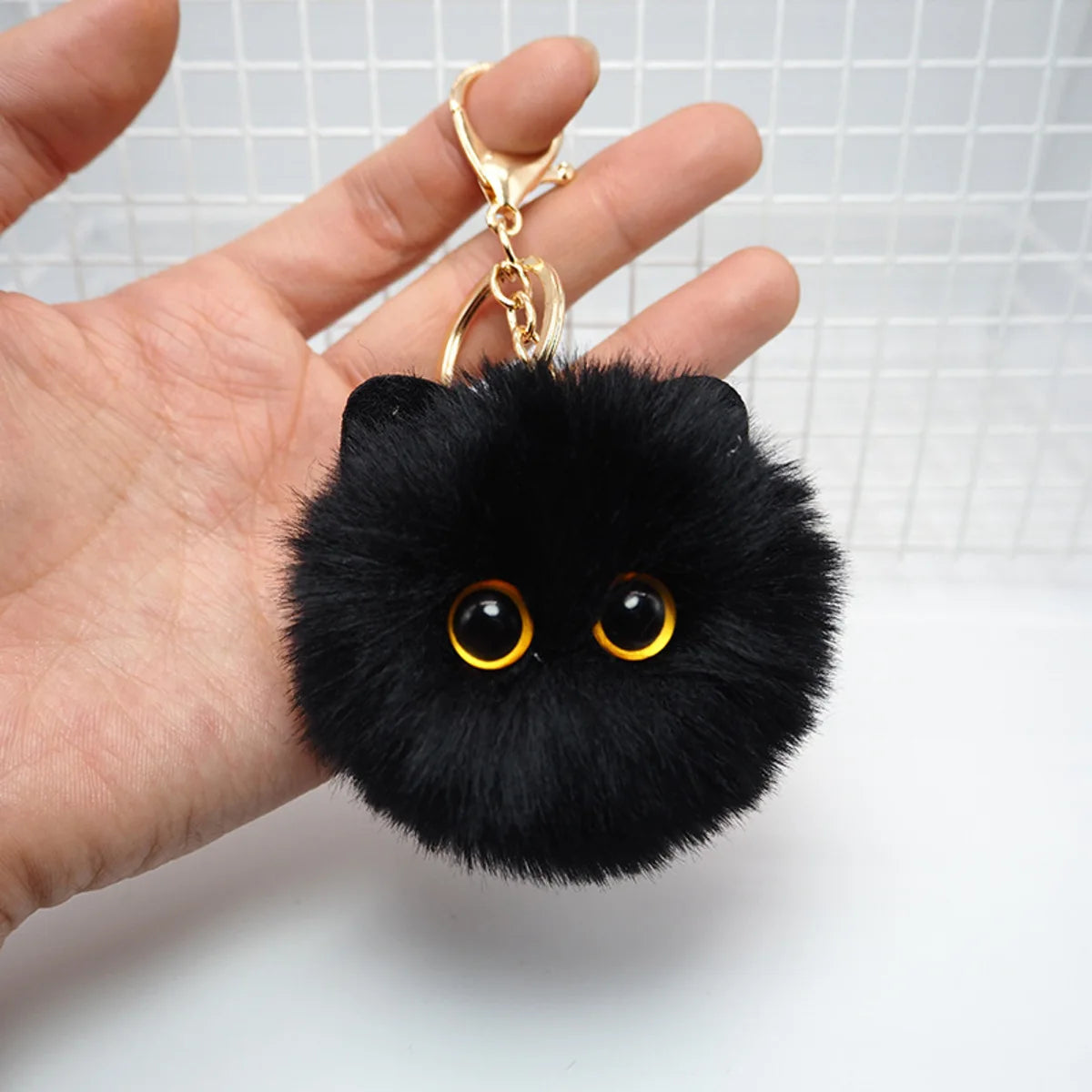 Black fluffy keychain with gold eyes held by a hand against a white background