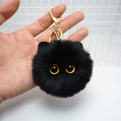 Black fluffy keychain with gold eyes held by a hand against a white background