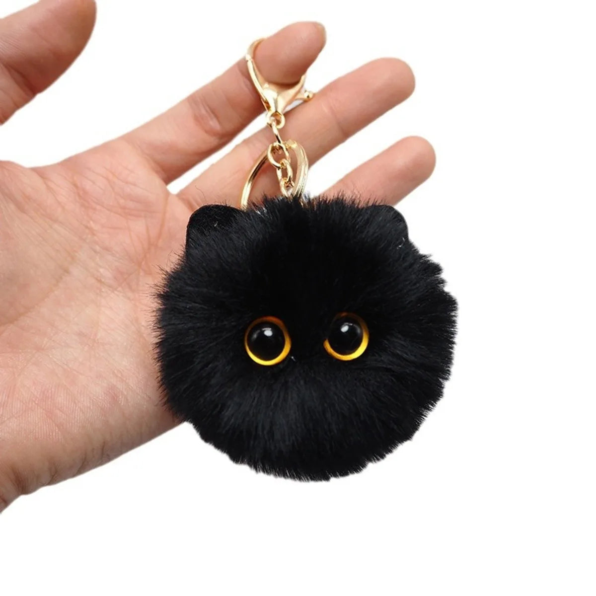 Black fluffy keychain with gold eyes held in a hand against a white background