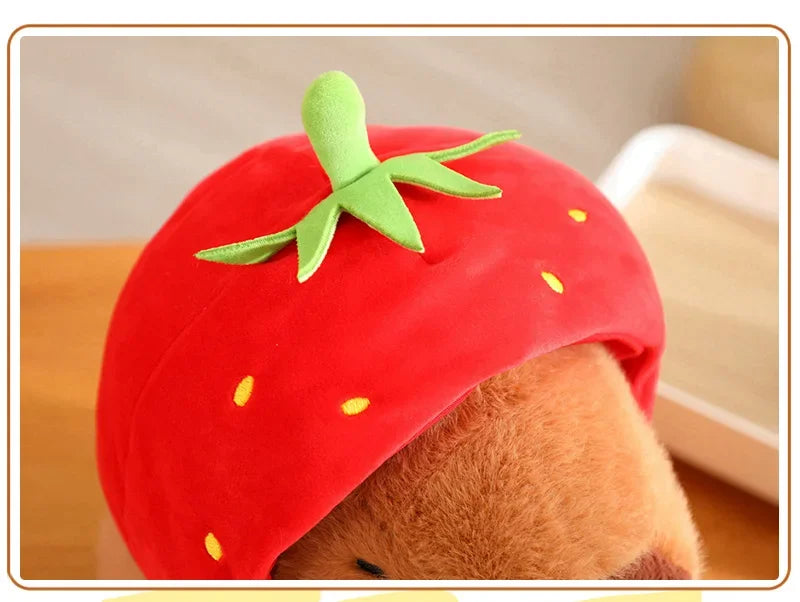 Teddy bear wearing a red strawberry-shaped hat with green leaves and yellow seeds on a wooden surface.