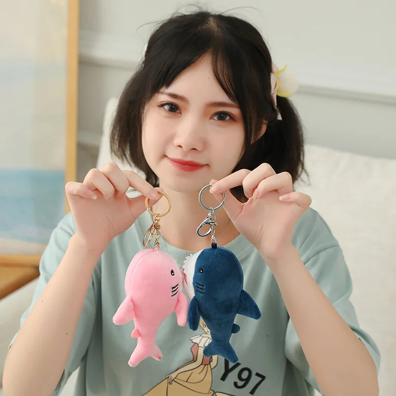 Person holding two plush shark keychains, one pink and one blue, in front of a blurred indoor background.
