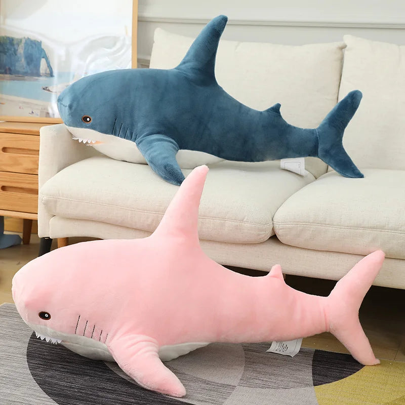 Two shark-shaped plush toys, one blue and one pink, on a couch and rug.