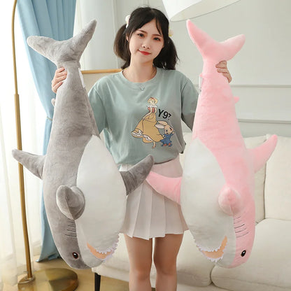 Woman holding a large gray shark plush toy and a pink shark plush toy in a room with a light blue curtain.