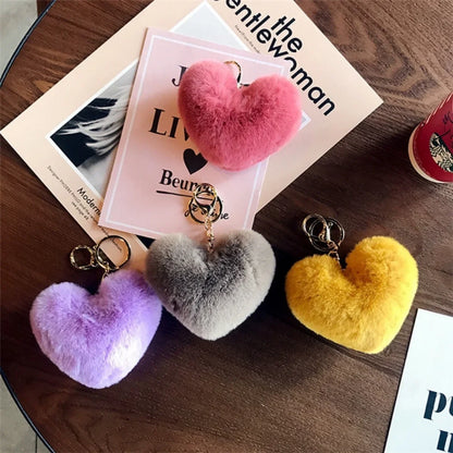 Heart-shaped fuzzy keychains in pink, gray, and yellow on a wooden surface with a magazine underneath.