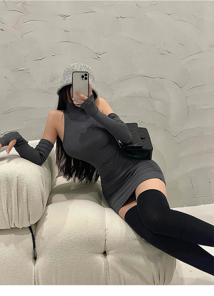 Person wearing a black outfit with long sleeves and gloves, sitting on a beige couch against a textured gray wall.