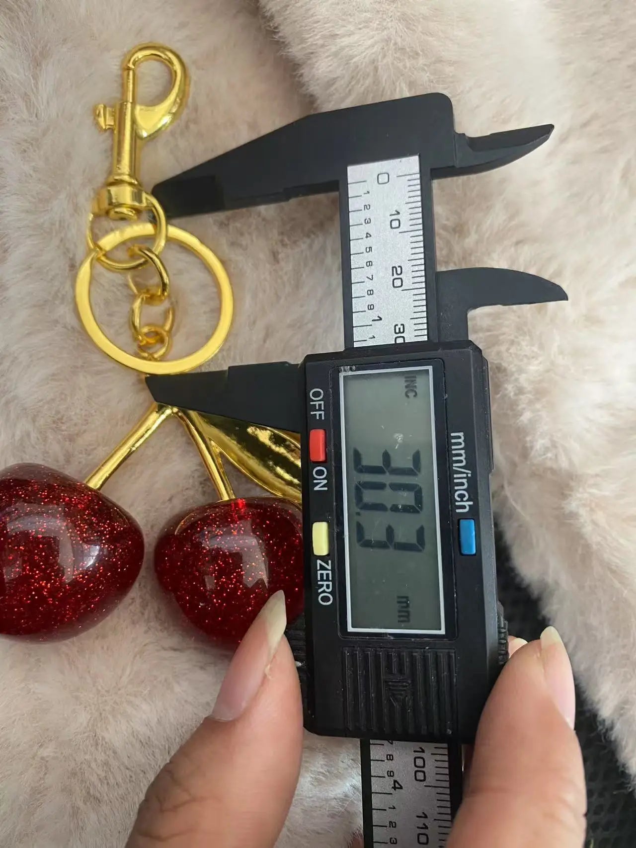Keychain with cherries and gold chain being measured with a digital caliper on a textured surface.
